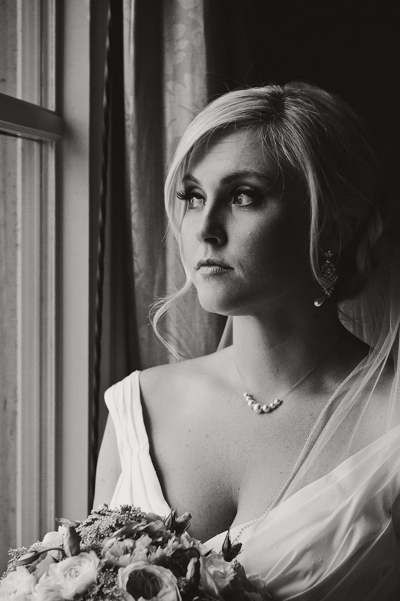 A black and white photo of a bride looking out of a window holding a bouquet of flowers.