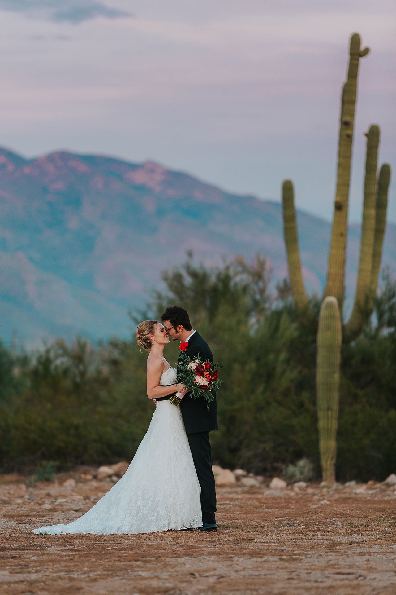 A bride and groom are kissing in front of a saguaro cactus in the desert.