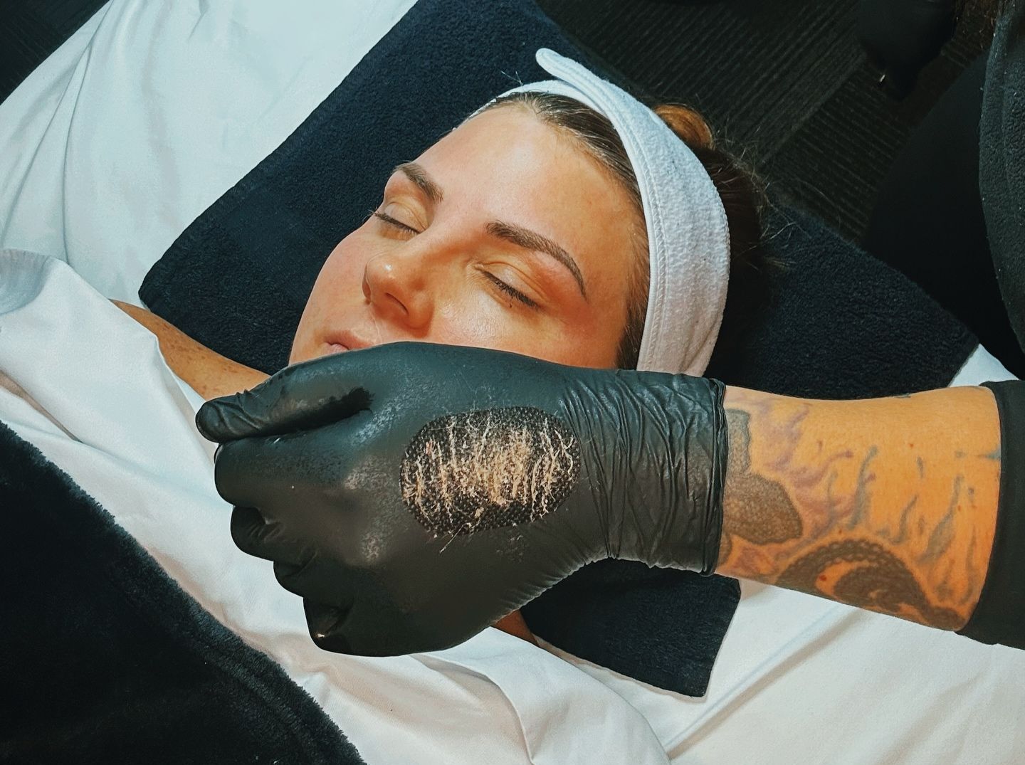 A woman is laying on a bed with her eyes closed while a person wearing black gloves holds her face.