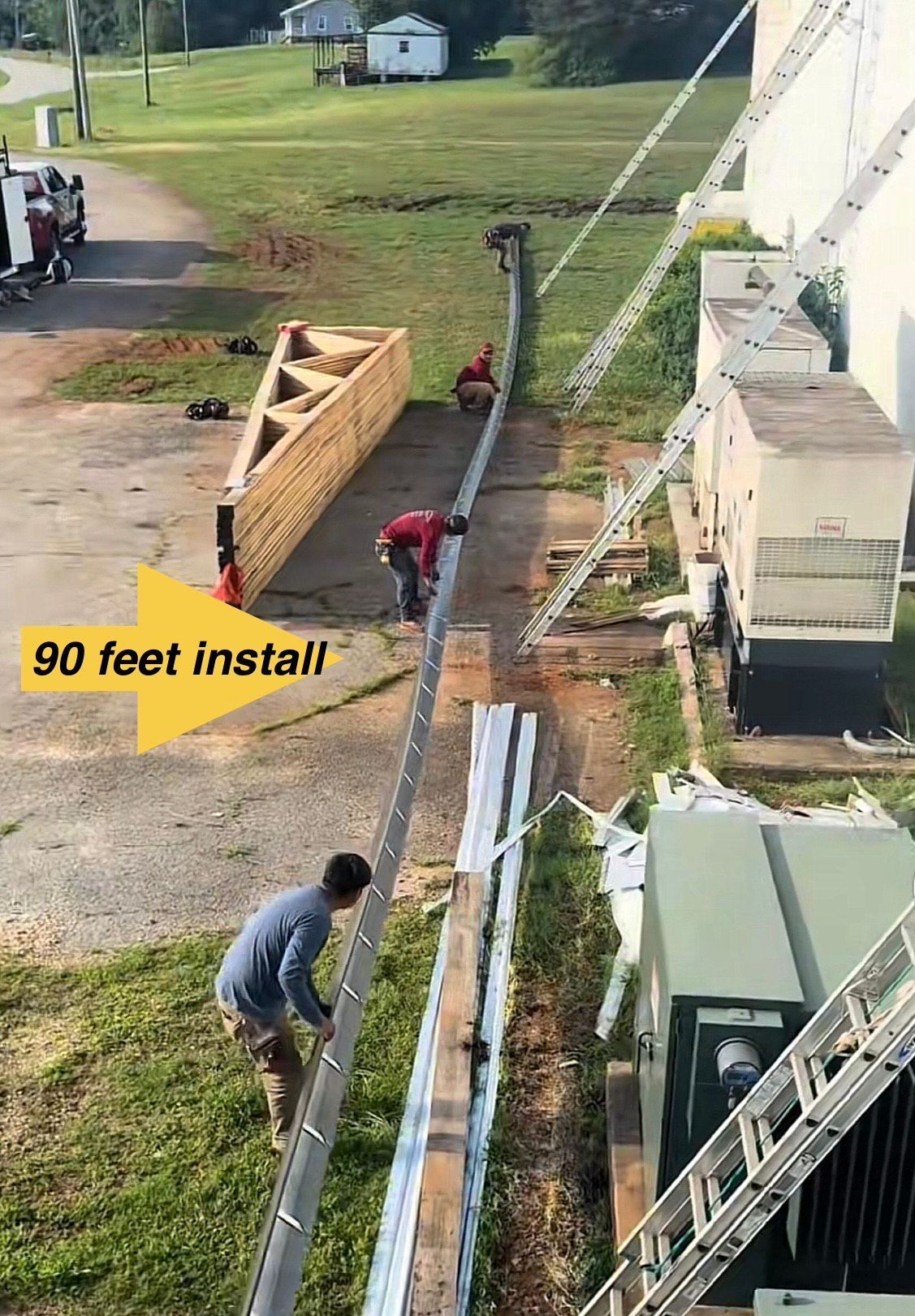 Construction workers install a 90-foot long metal piece along a building, using ladders.