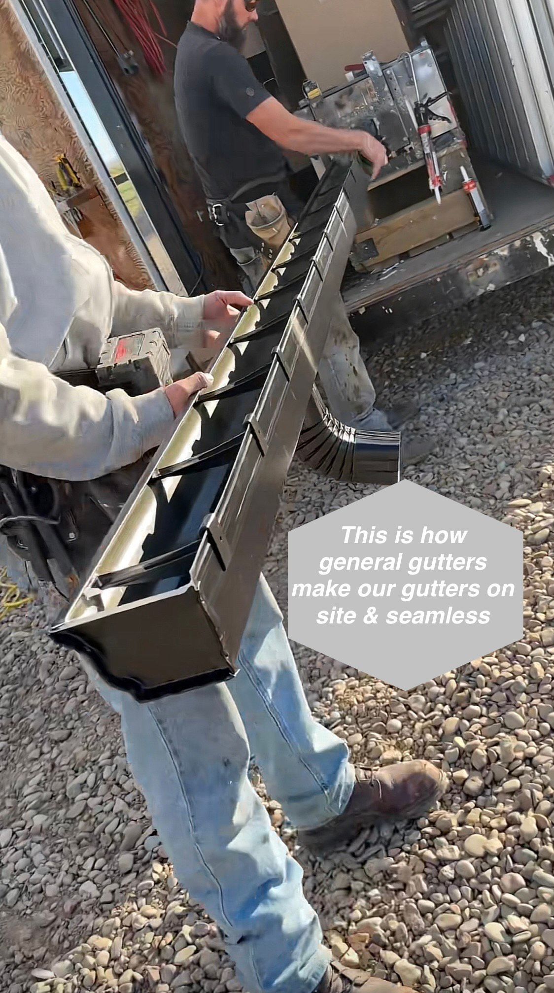 Two workers installing a long, grey gutter. One holds the gutter; the other stands nearby.