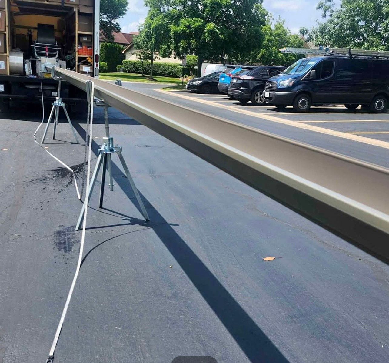 A long, brown gutter held by tripods on asphalt, next to a truck. Parked cars in background.