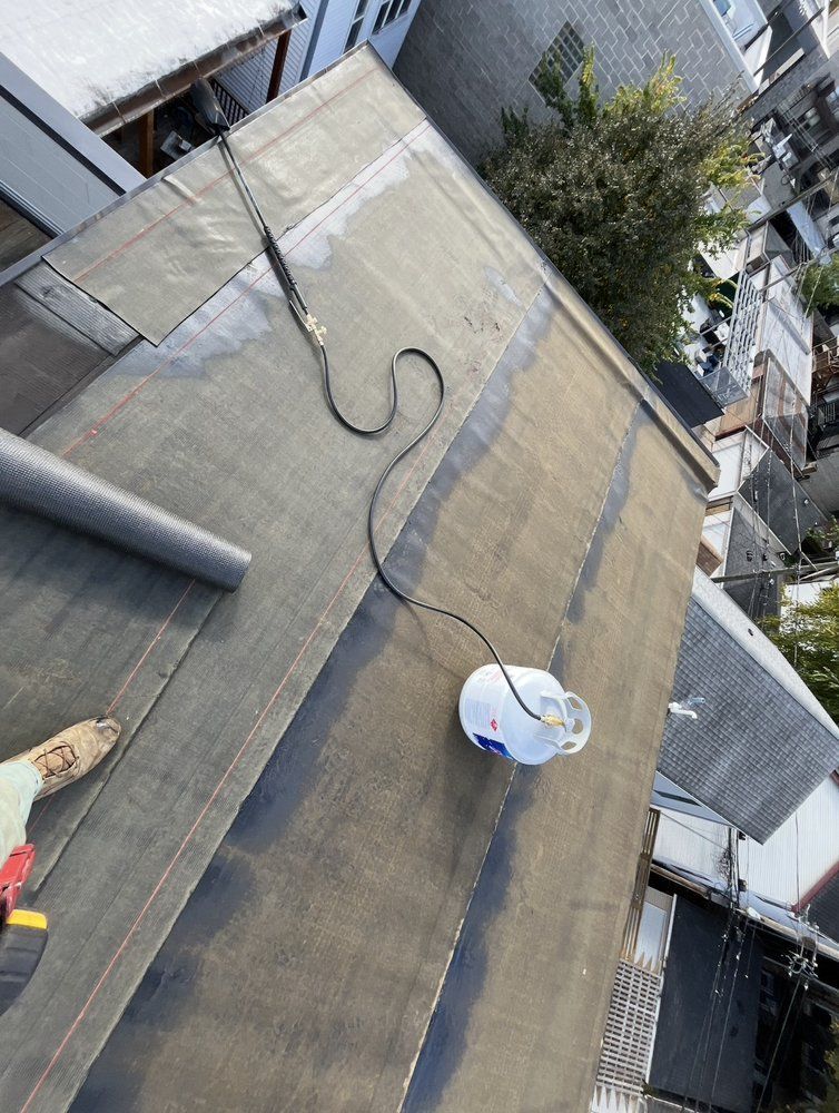 Roofer working on a flat roof. Propane tank and hose visible. Gray and black roofing material.