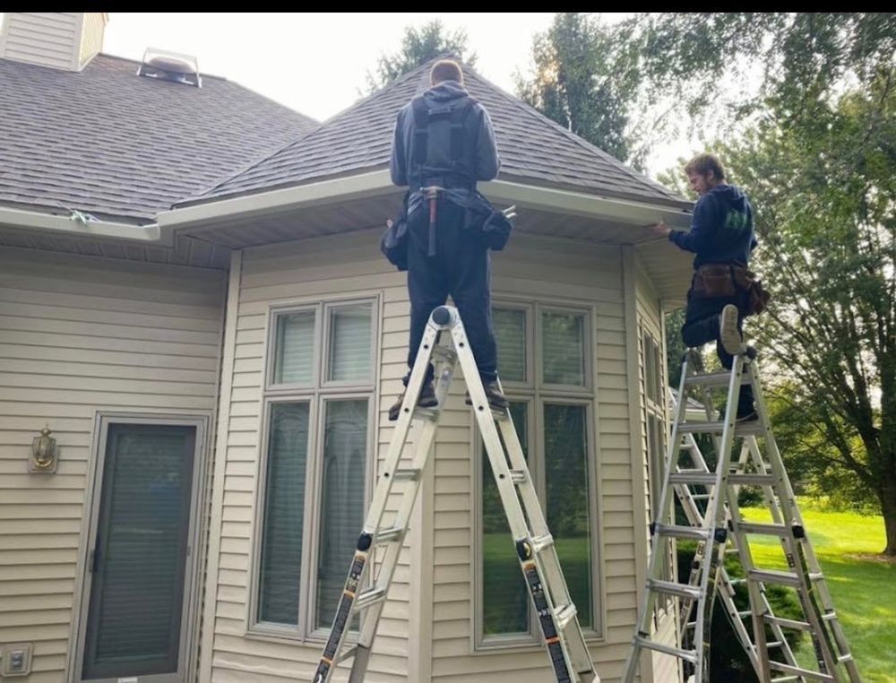 Two workers on ladders cleaning gutters of a light-colored house. Green trees and grass visible.