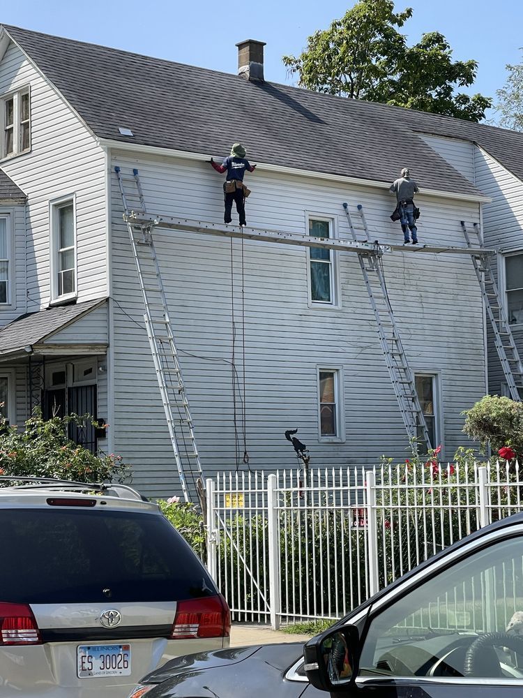 Two people on a house roof using ladders. A white house with multiple windows and a car parked in front.