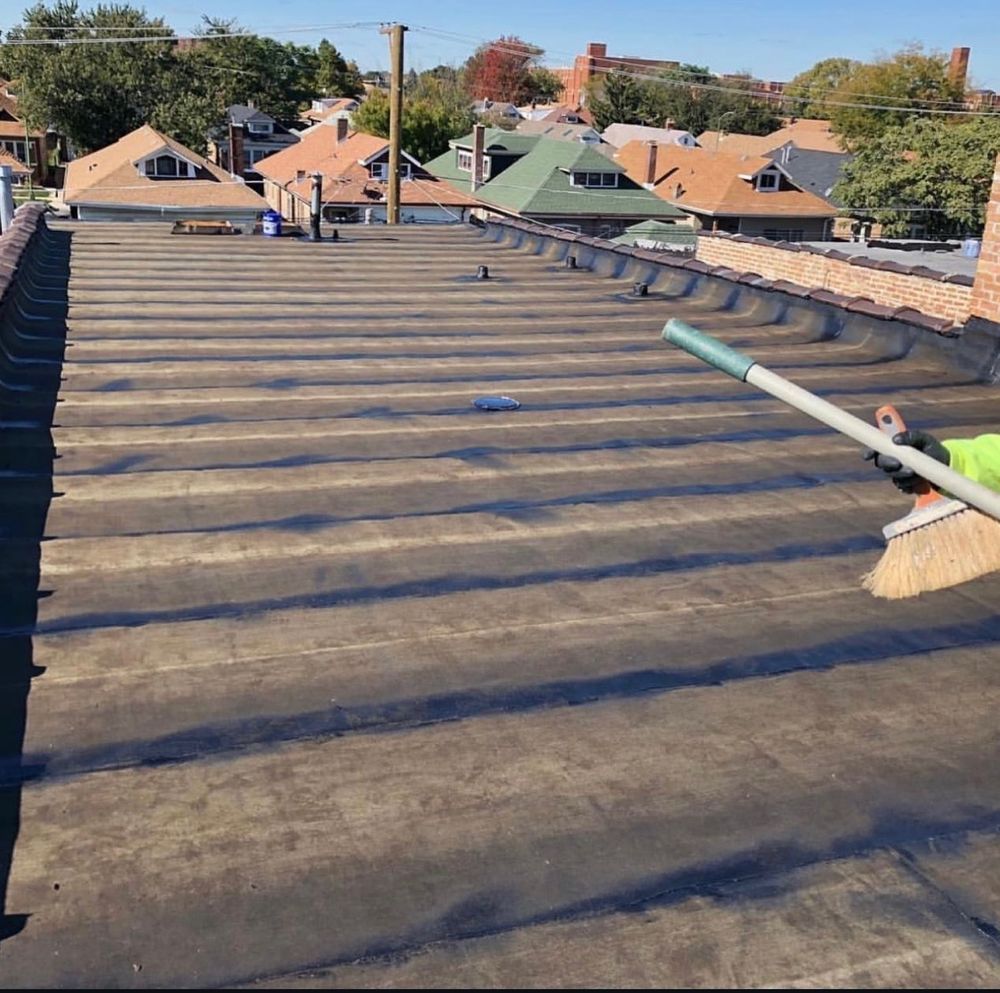 Person cleaning a flat, black roof with a long-handled brush on a sunny day.