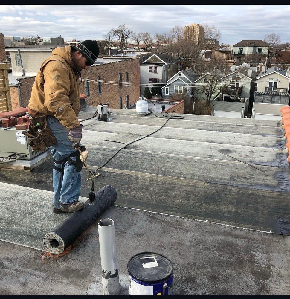 Roofer rolls out roofing material on a flat roof. He's connected to a safety line. Buildings in the background.