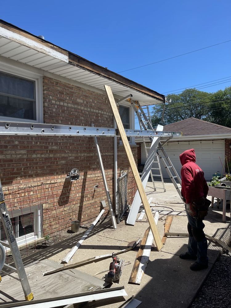 Construction worker on a concrete patio next to a brick house, using ladders to work on the roofline.