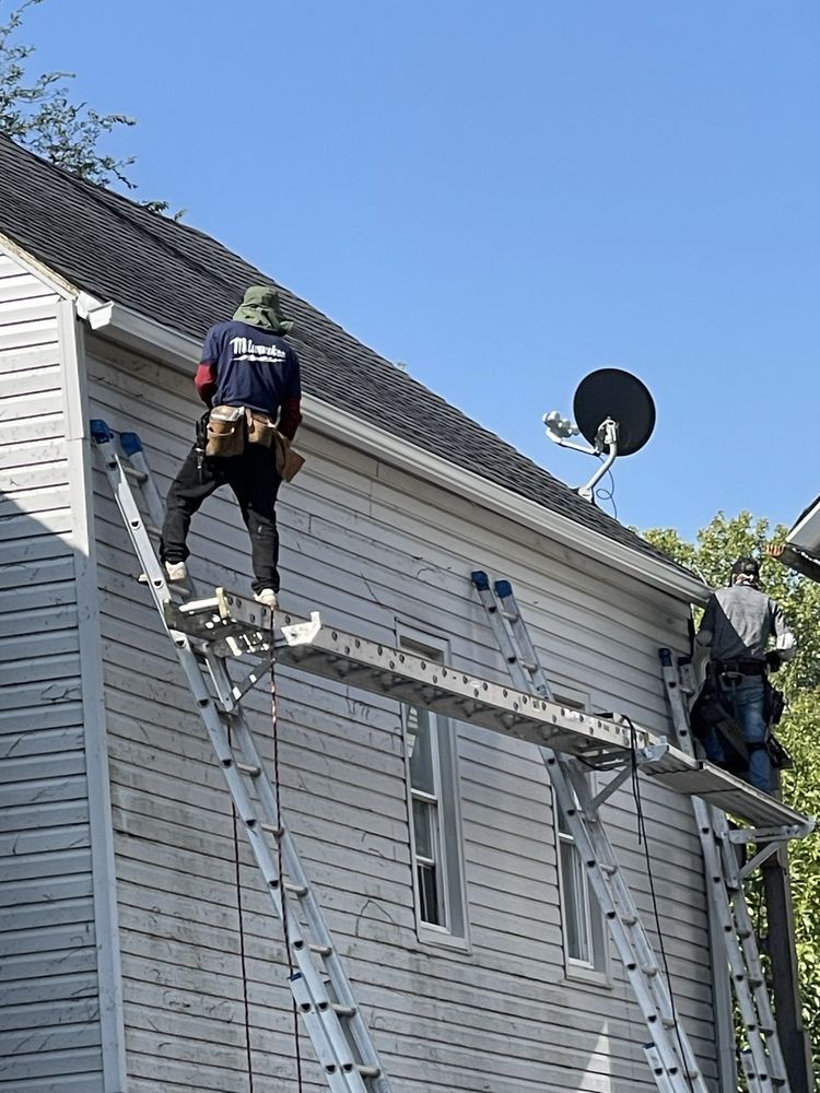 Two workers on ladders accessing a house's siding and gutters. One stands on a scaffold. Clear blue sky.