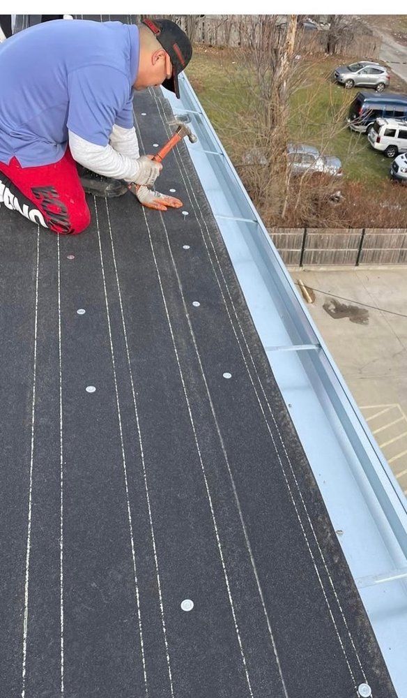 Person kneeling on a flat roof, hammering. The roof is black with silver metal trim.