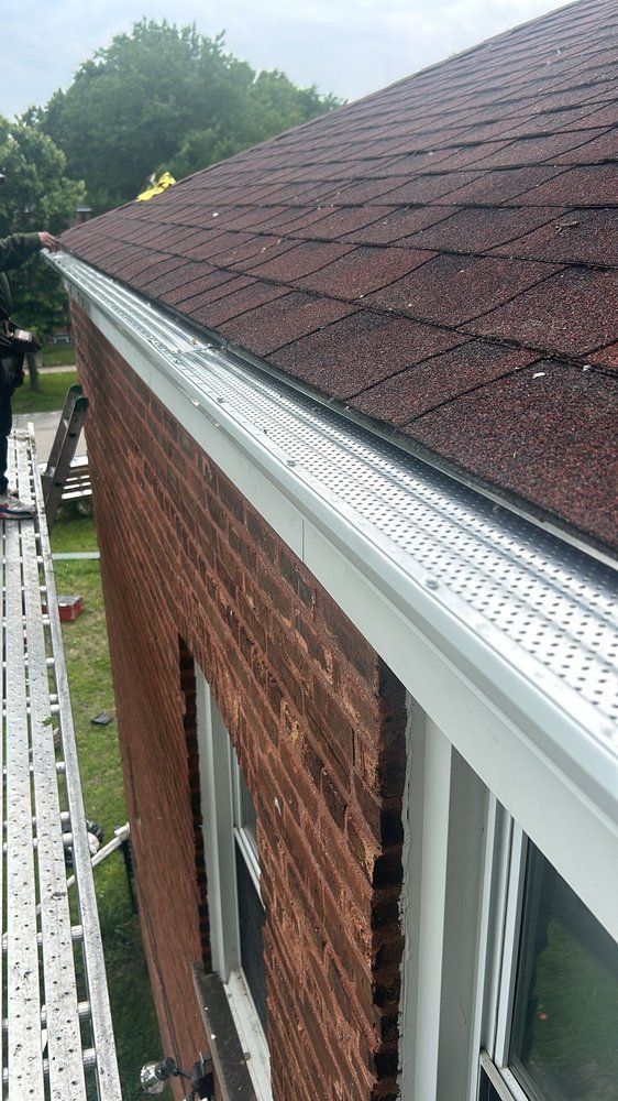 Gutter with mesh cover on a brick building with a red shingle roof. A ladder sits nearby.