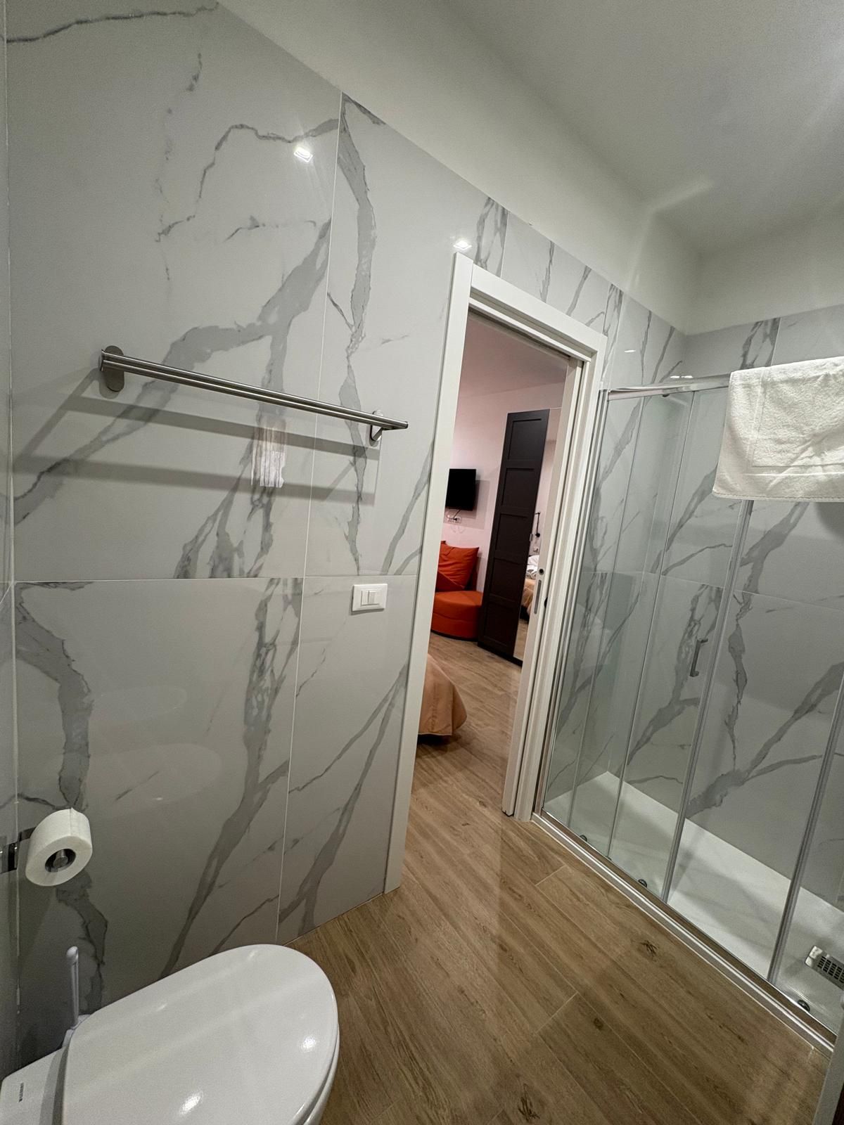 Bagno con piastrelle effetto marmo, portasciugamani, WC e una porta aperta che conduce a una zona soggiorno con divano arancione.