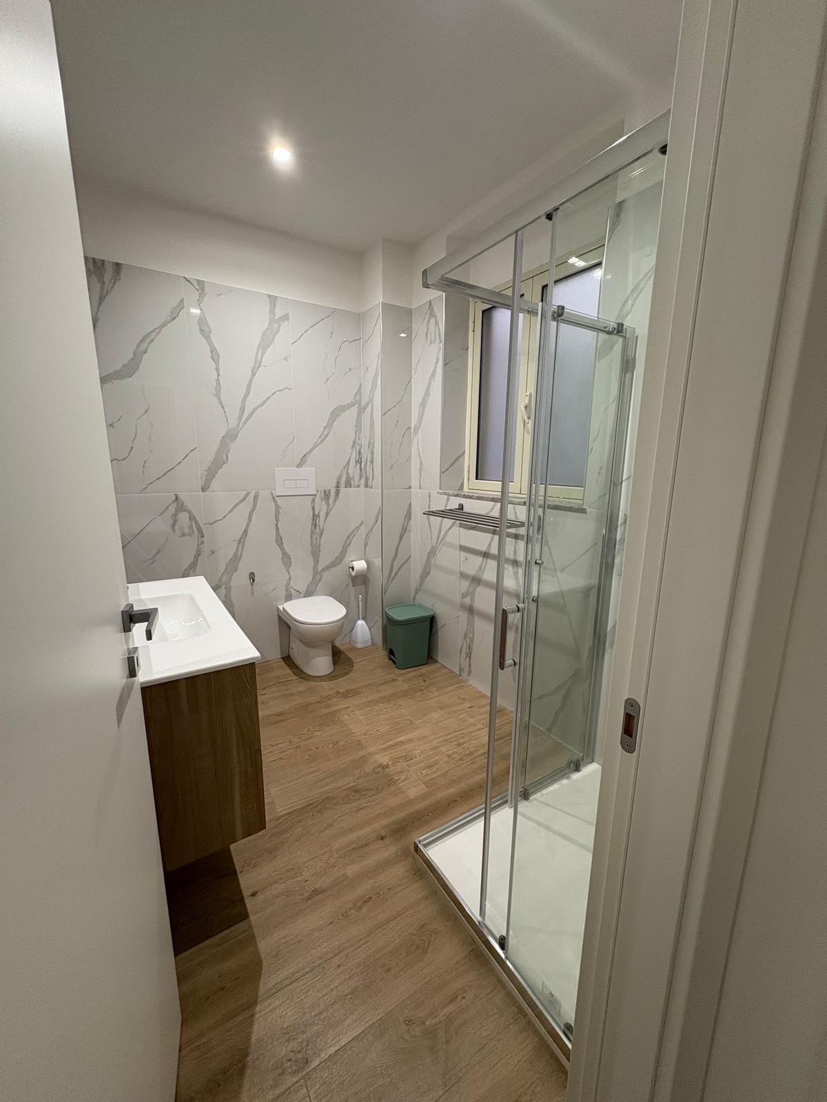 Bagno con piastrelle effetto marmo, lavabo in legno, doccia in vetro, WC e pavimento effetto legno.