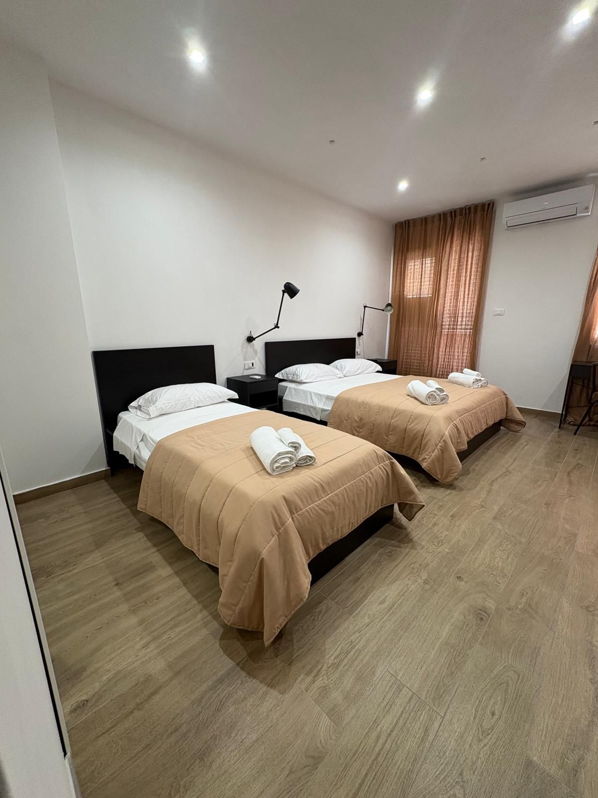 Due letti singoli in una stanza luminosa con lenzuola marrone chiaro, asciugamani bianchi e testiere nere.