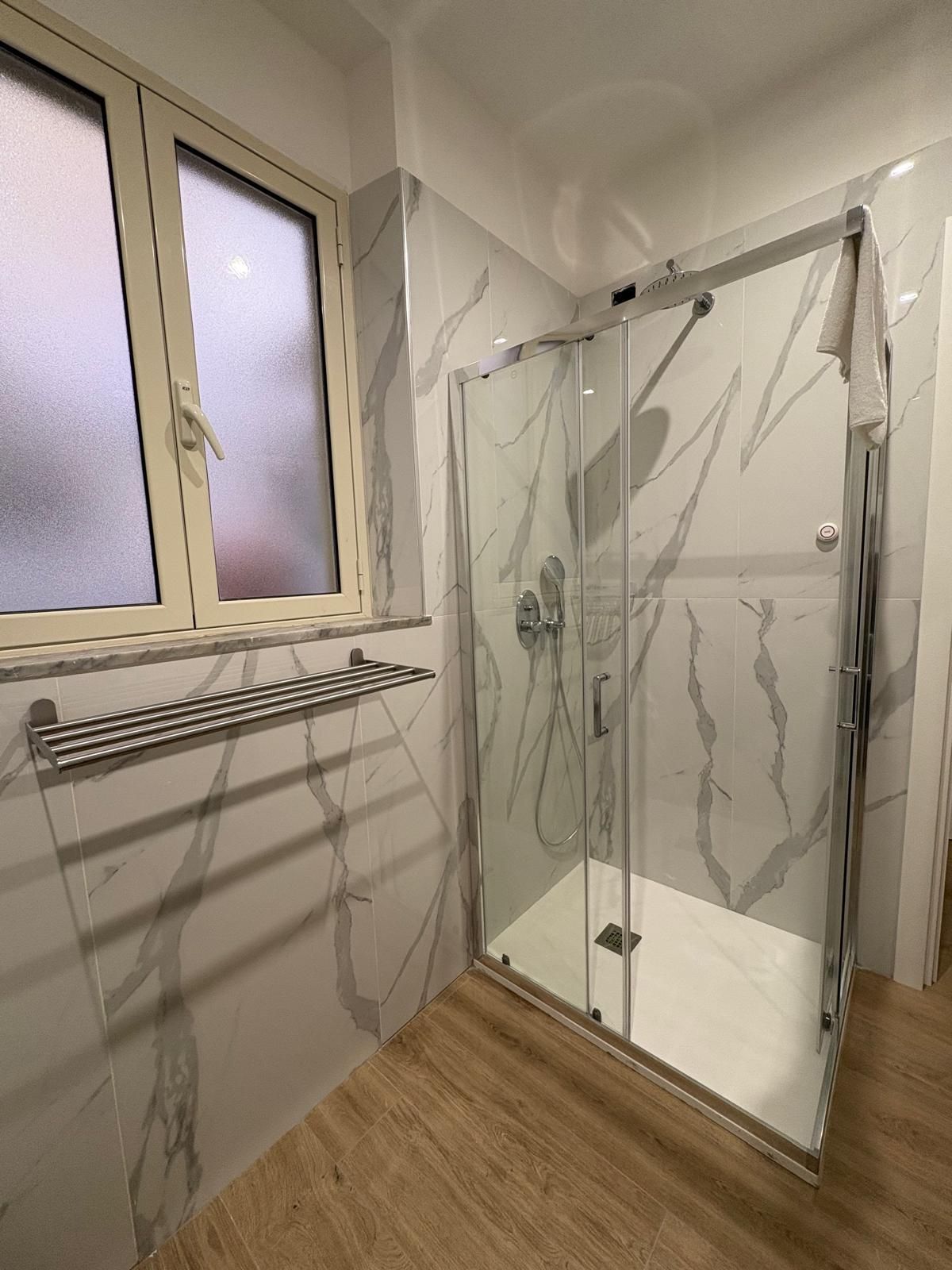 Un bagno con doccia in vetro, pareti in marmo bianco, finestra e portasciugamani.