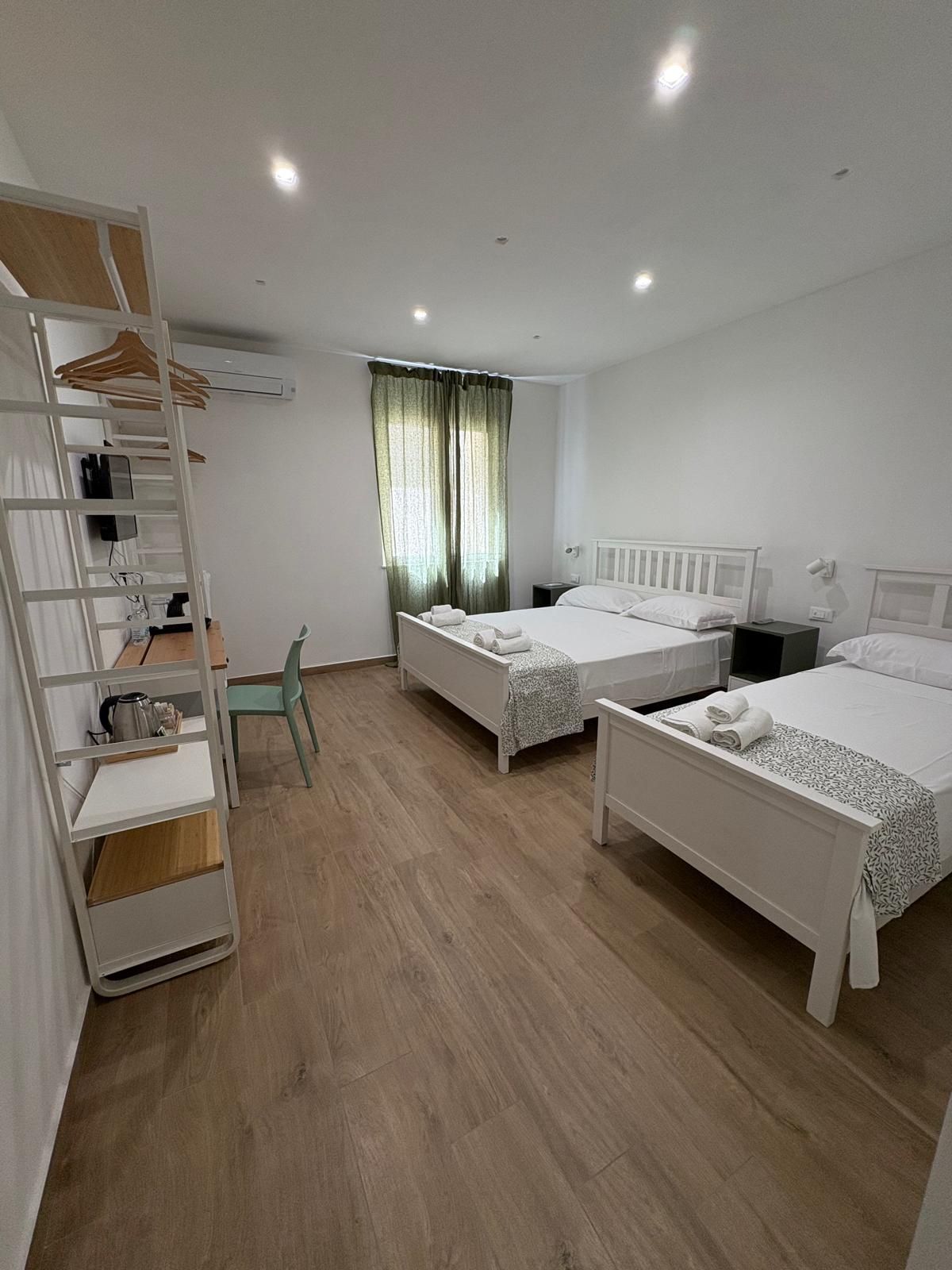 Luminosa camera d'albergo con due letti bianchi, una scrivania e uno scaffale alto.