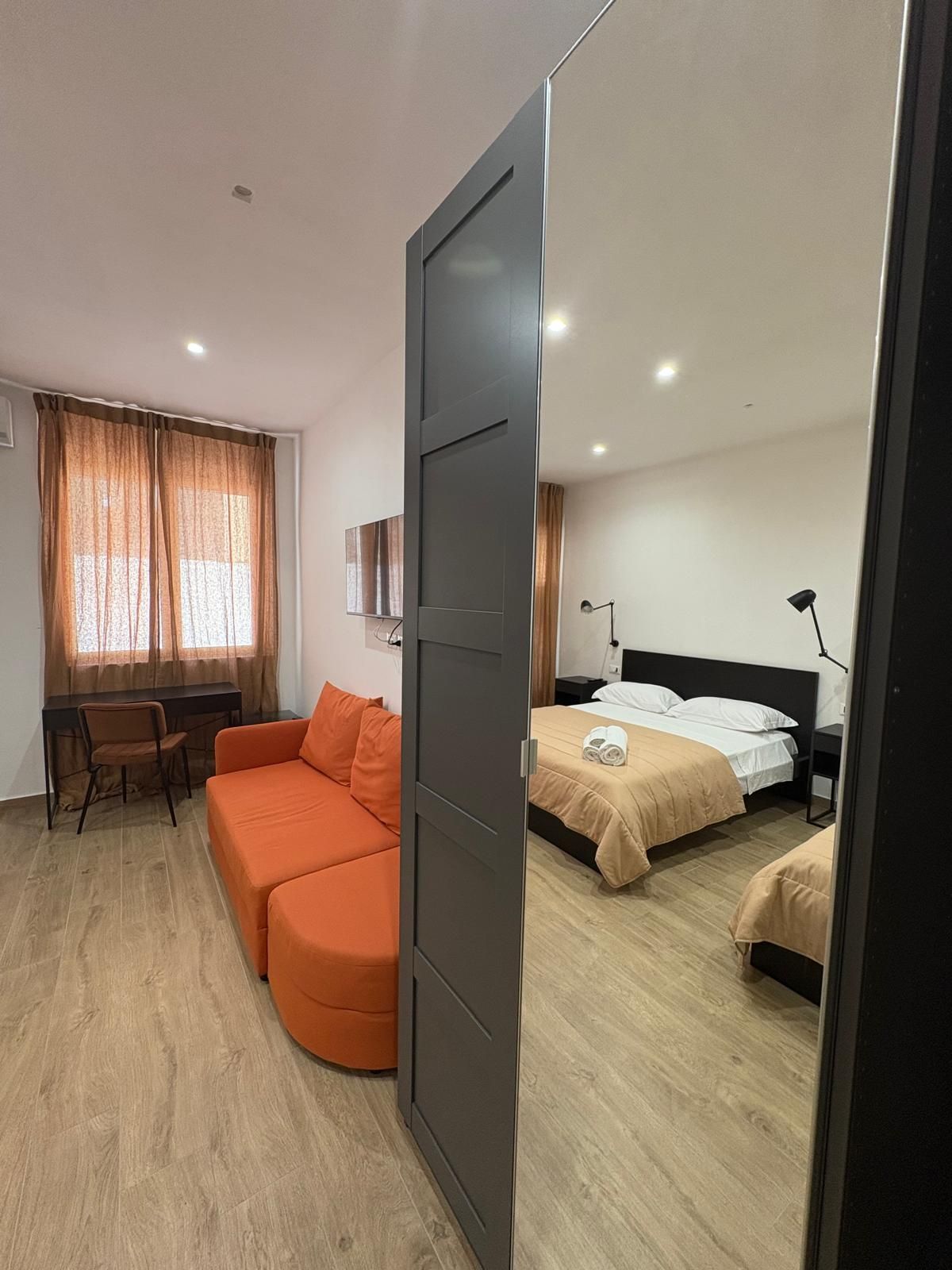 Specchio che riflette una camera da letto e una parte del soggiorno. Divano arancione, letto con piumone color cuoio, tende marroni e pavimento in legno.