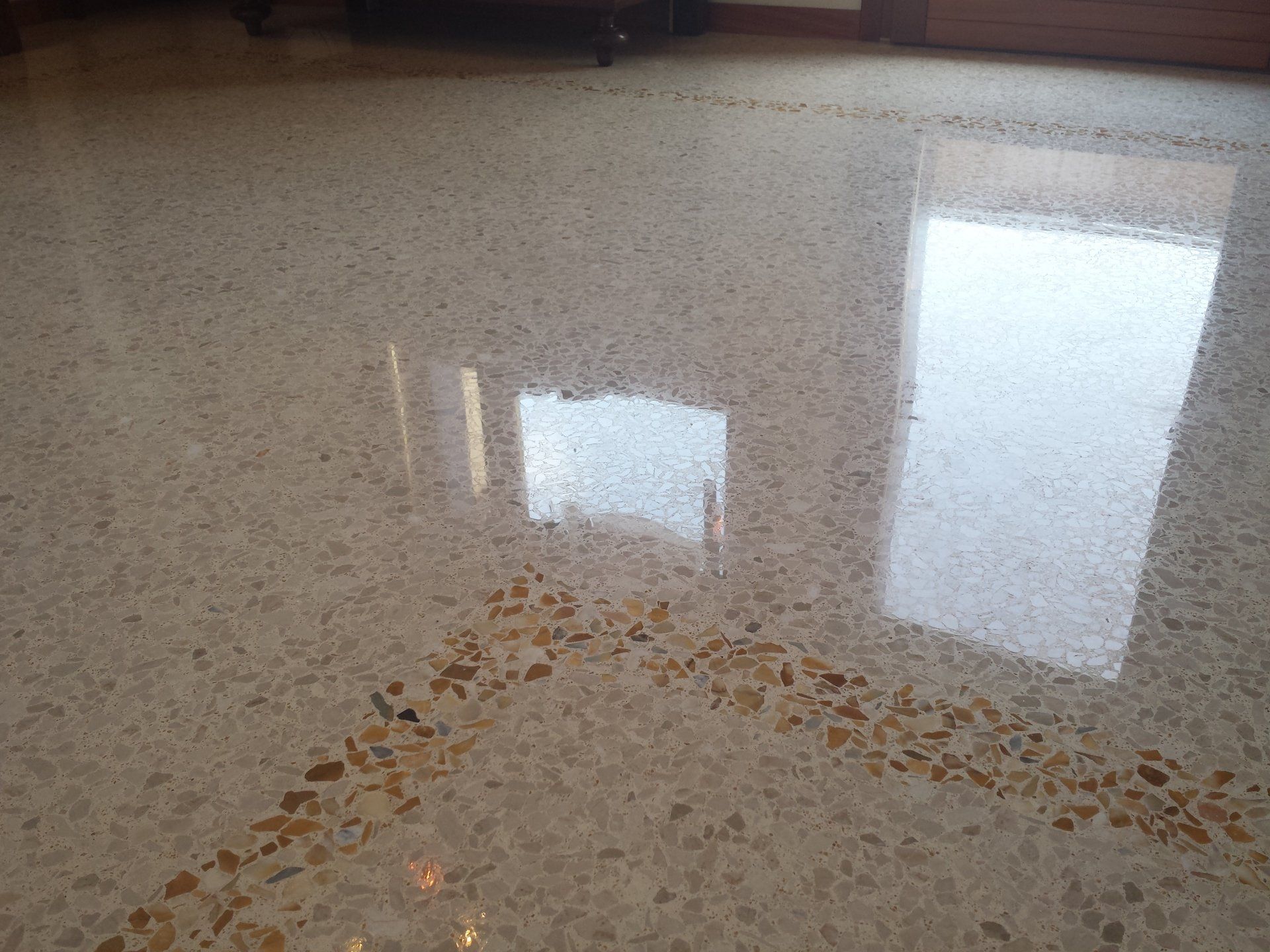 Pavimento in terrazzo lucidato con piccole scaglie di aggregati color crema e marrone, che riflettono la luce intensa proveniente dalle finestre vicine.