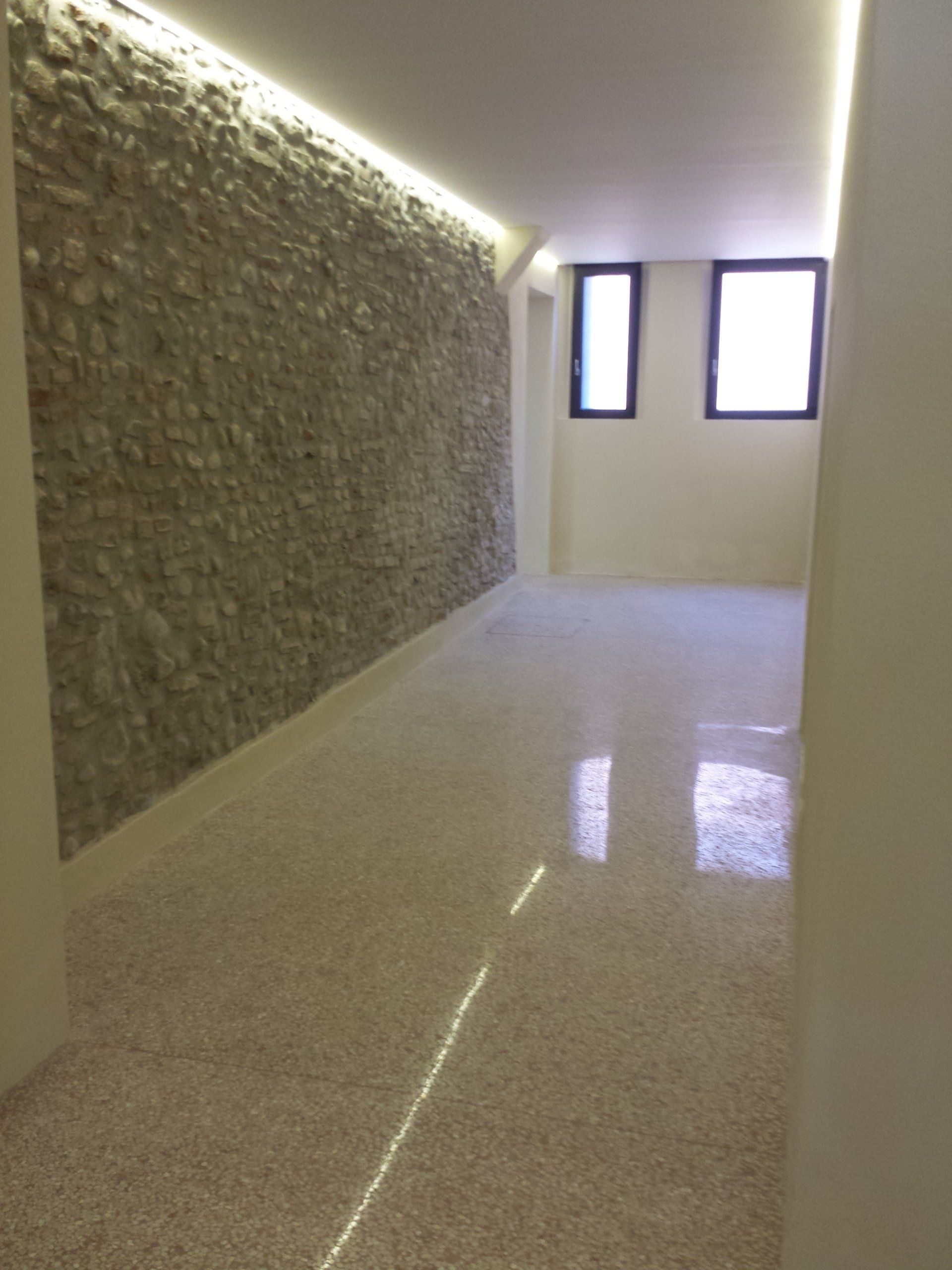 Un lungo corridoio con una parete rustica con texture in pietra sulla sinistra, un pavimento liscio e chiaro e due finestre all'estremità più lontana.