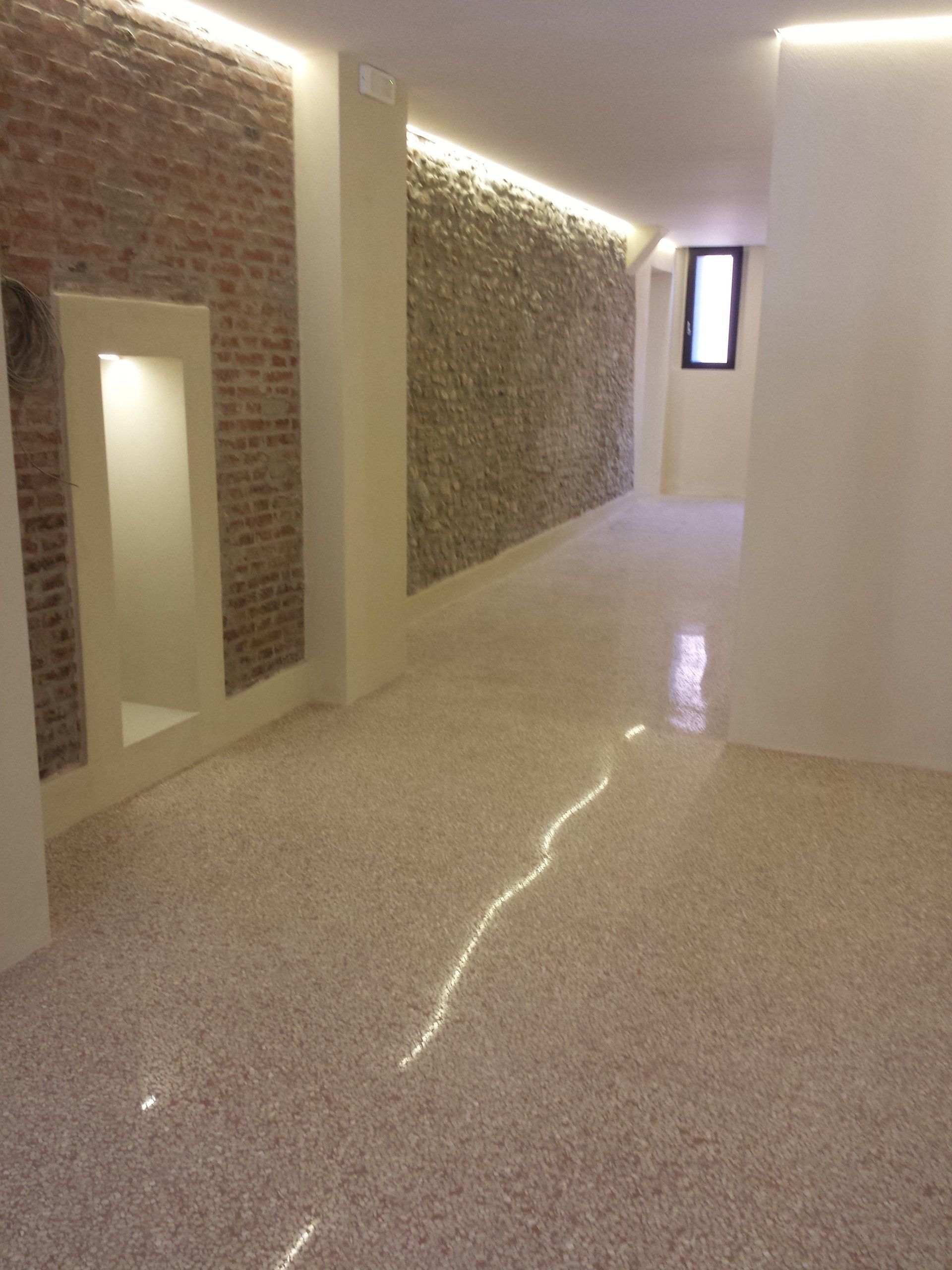Un corridoio interno con pavimento in terrazzo lucidato, pareti in mattoni e pietra a vista e illuminazione a incasso.