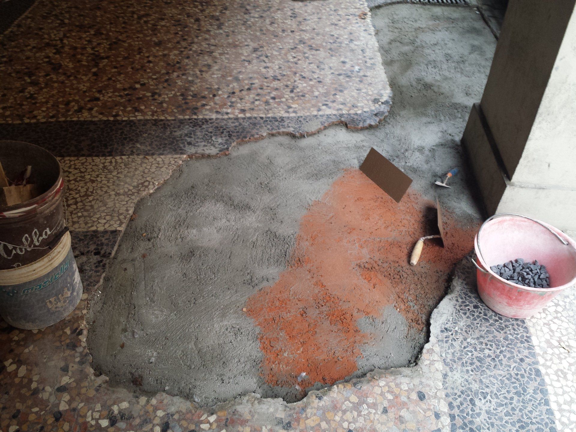 Una porzione di cemento grigio fresco riparato su un pavimento in pietra ruvida, con una cazzuola, un secchio e un contenitore nelle vicinanze.