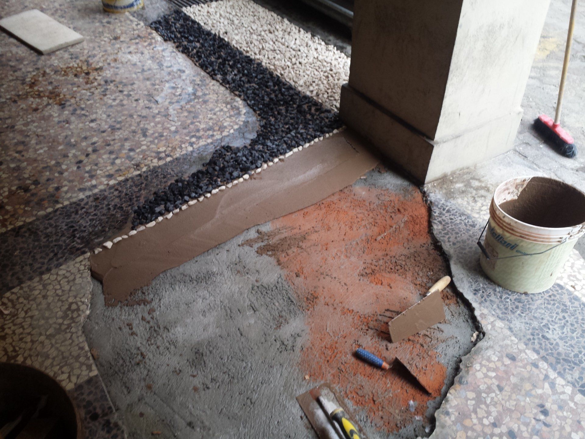 Un cantiere edile mostra un pavimento parzialmente riempito con pietre bianche e nere accanto a cemento e cazzuole.
