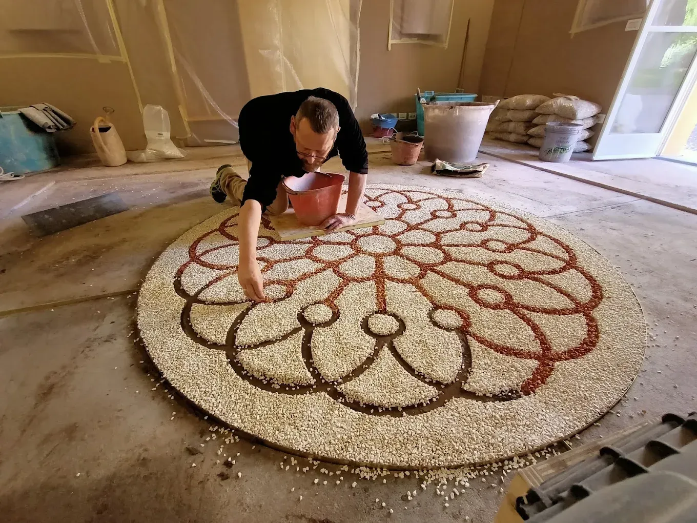 Una persona inginocchiata su un pavimento di cemento, sistema con cura ciottoli bianchi e rosso-marroni in un intricato disegno a mandala.