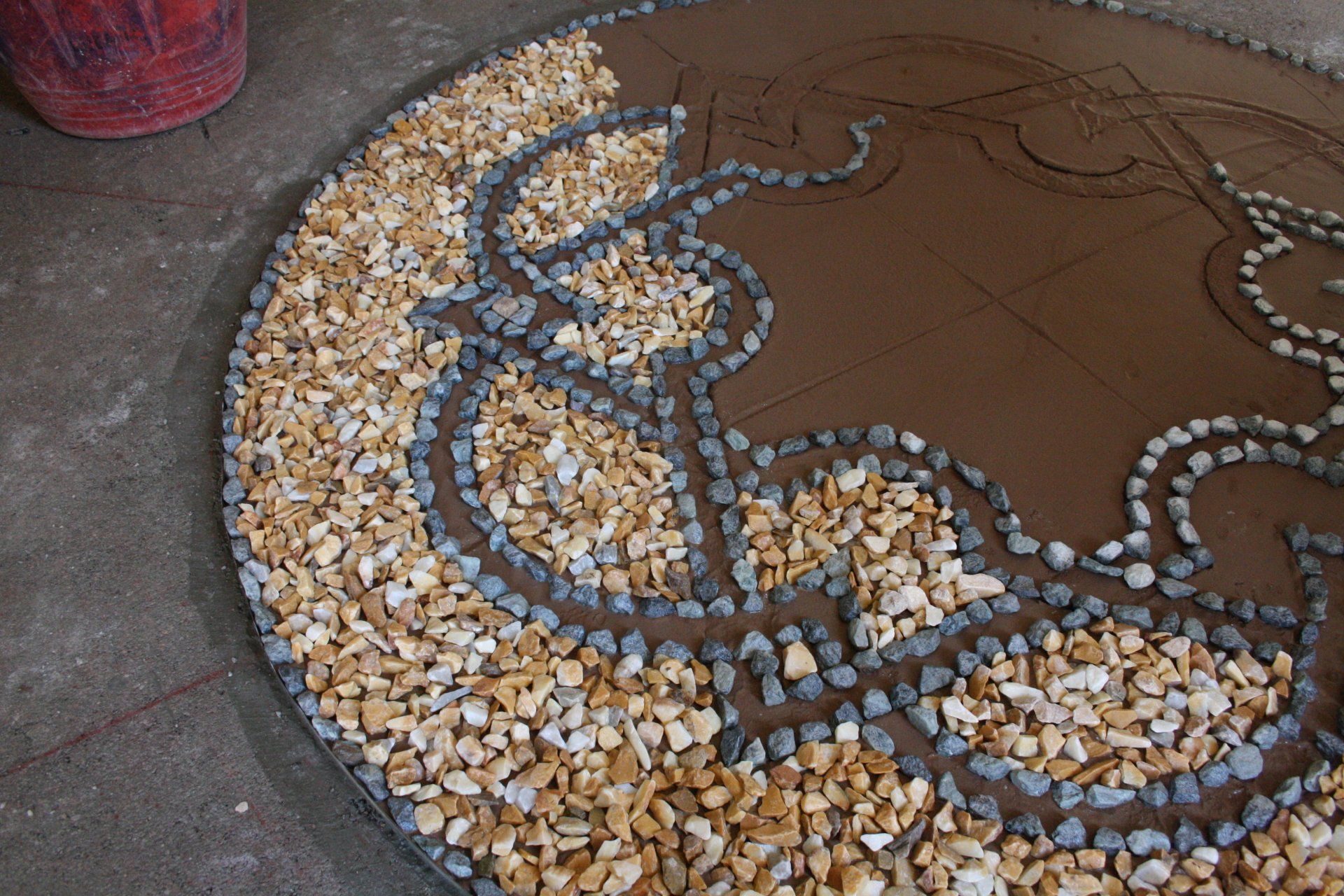 Un mosaico circolare in lavorazione, caratterizzato da un motivo di bordo in pietra scura riempito con ghiaia chiara su una base marrone.