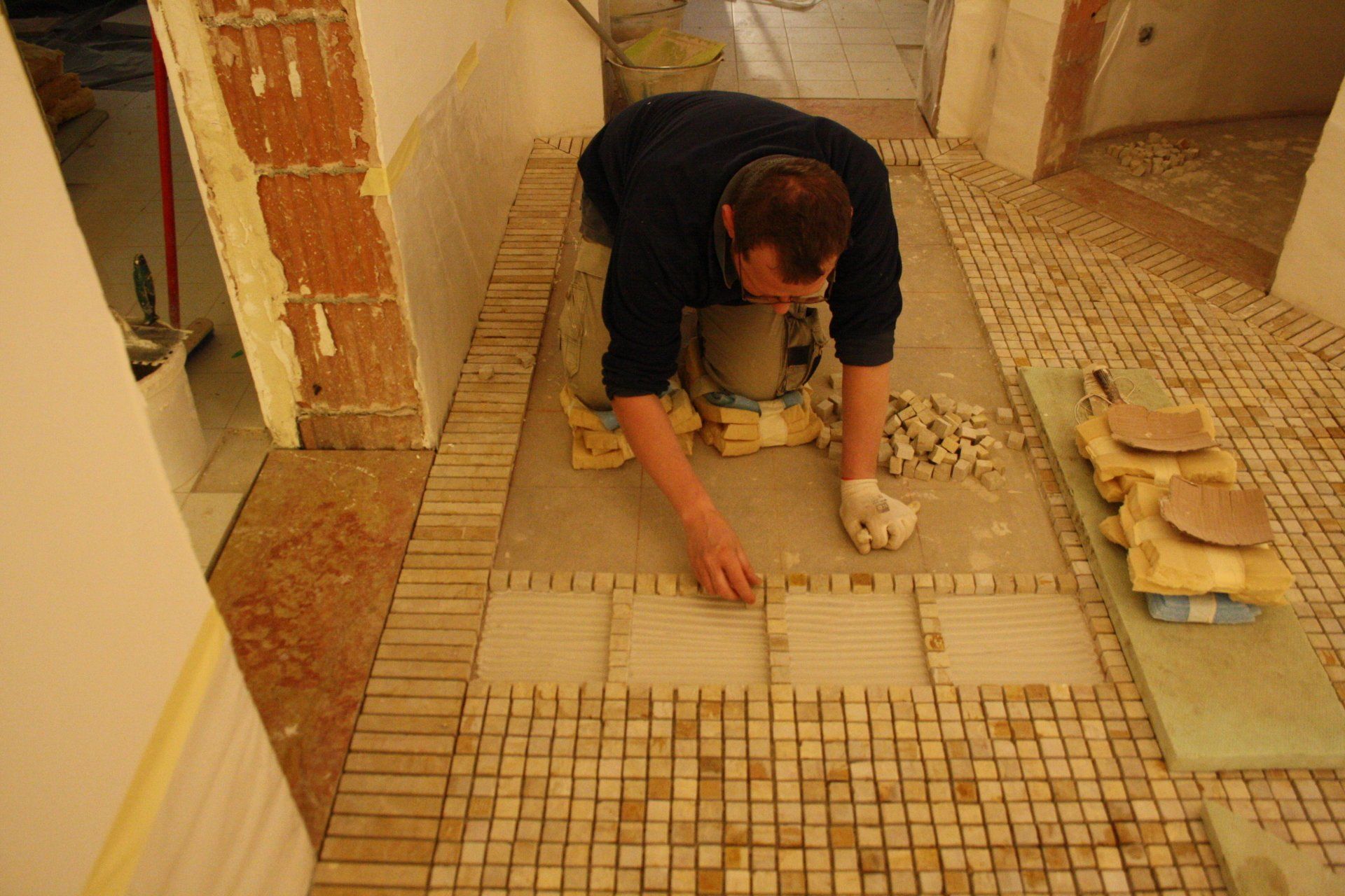 Un operaio è inginocchiato sul pavimento e sta sistemando con cura piccole tessere di mosaico di colore chiaro, formando una griglia in uno spazio interno.