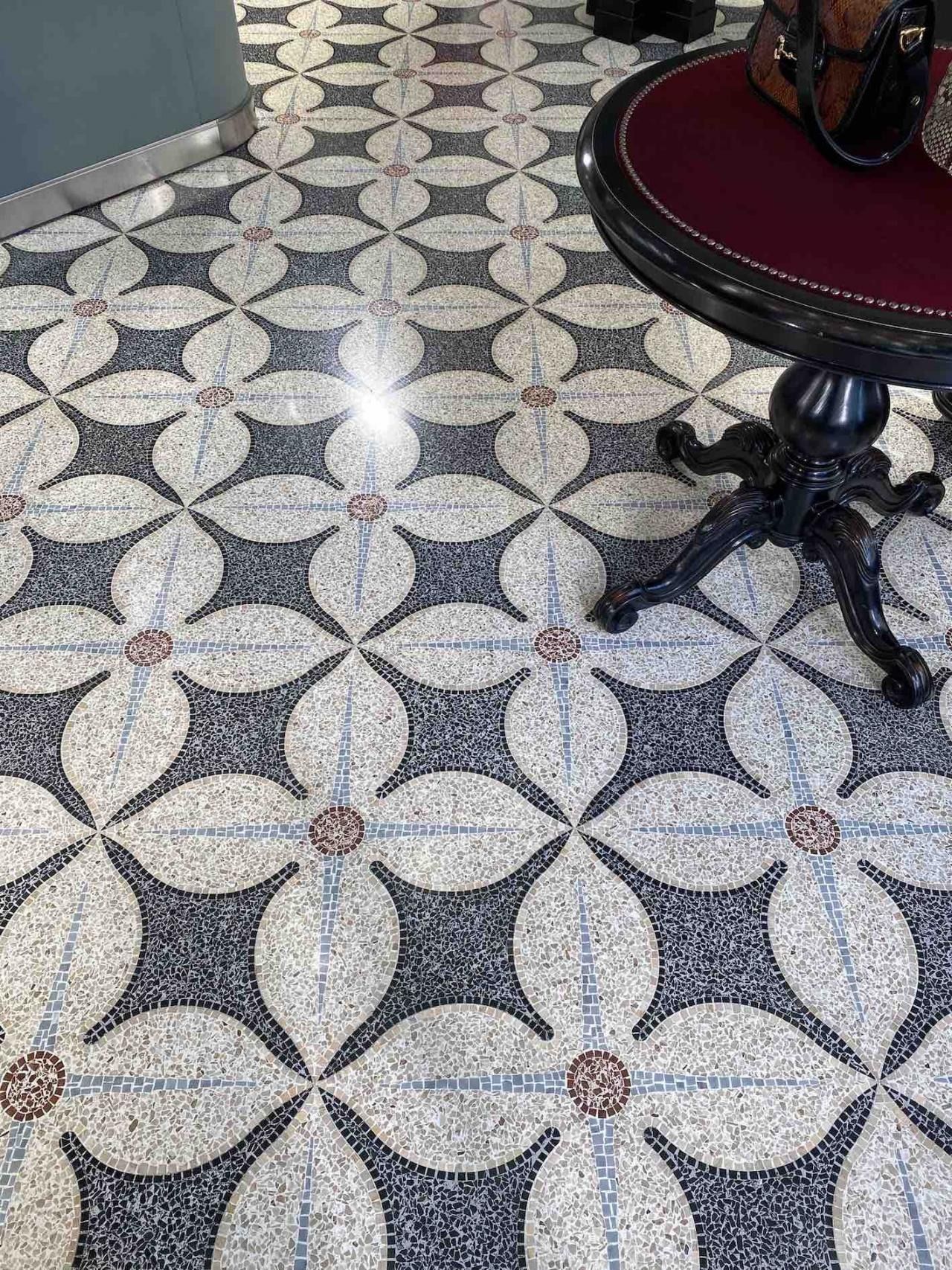 Un pavimento decorativo in piastrelle a mosaico con un motivo floreale ripetuto in bianco e nero, parzialmente nascosto da un tavolo rotondo.