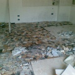 Una stanza in fase di ristrutturazione con un pavimento di piastrelle rotte, detriti sparsi per terra e pareti bianche e spoglie.