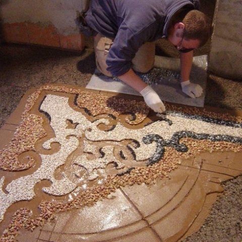 Una persona che indossa i guanti posiziona con cura piccole pietre colorate formando un mosaico decorativo sul pavimento.