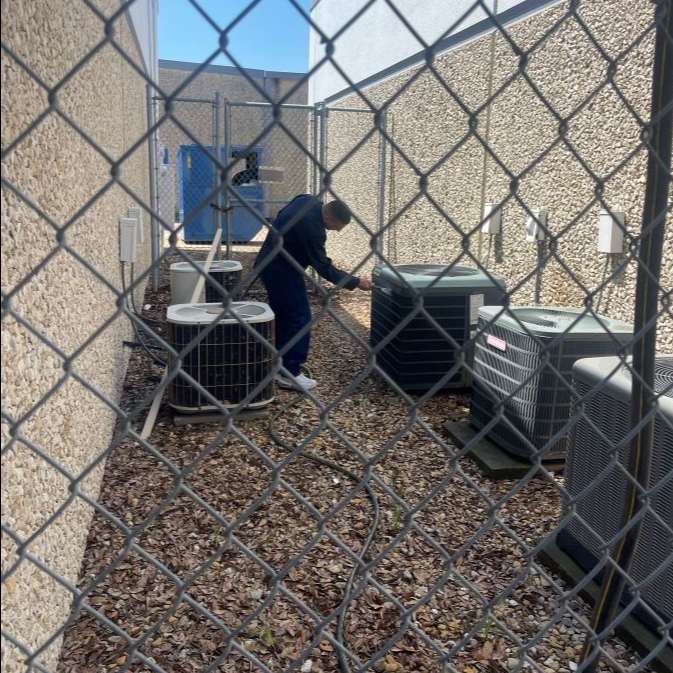 A man is working on an air conditioner outside of a building behind a chain link fence.