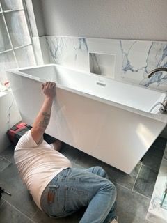 A man is kneeling on the floor next to a bathtub in a bathroom.