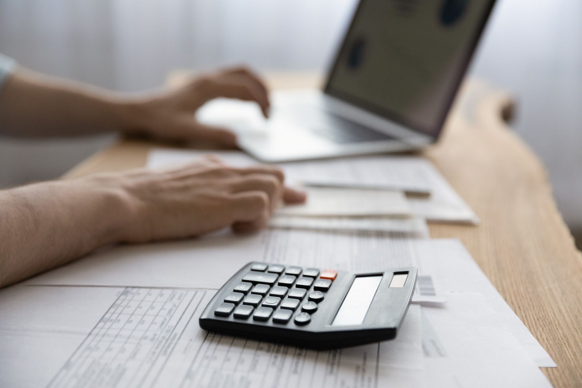 Person using laptop with a calculator and papers on a desk; financial calculations.