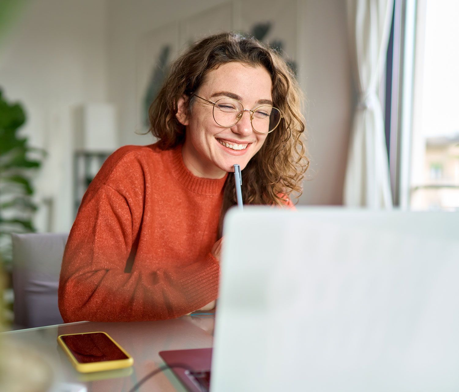 Woman wearing glasses smiles while looking at a laptop. Holding a pen, sitting at a desk indoors.
