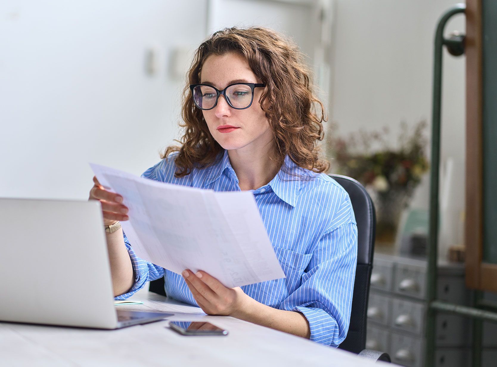 Woman with curly hair and glasses reviewing documents while using a laptop.