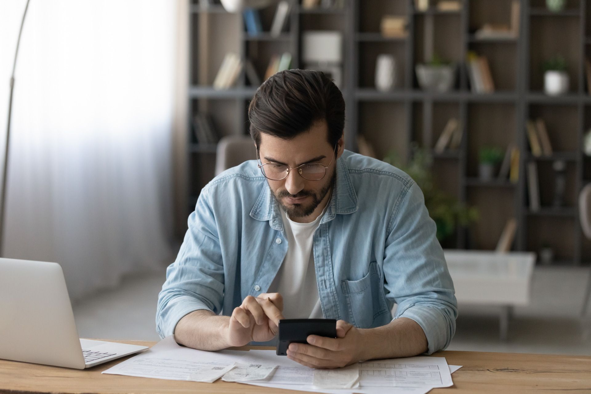 Man in blue shirt, glasses, using phone, sitting at a desk with laptop, documents, and a bookshelf in background.