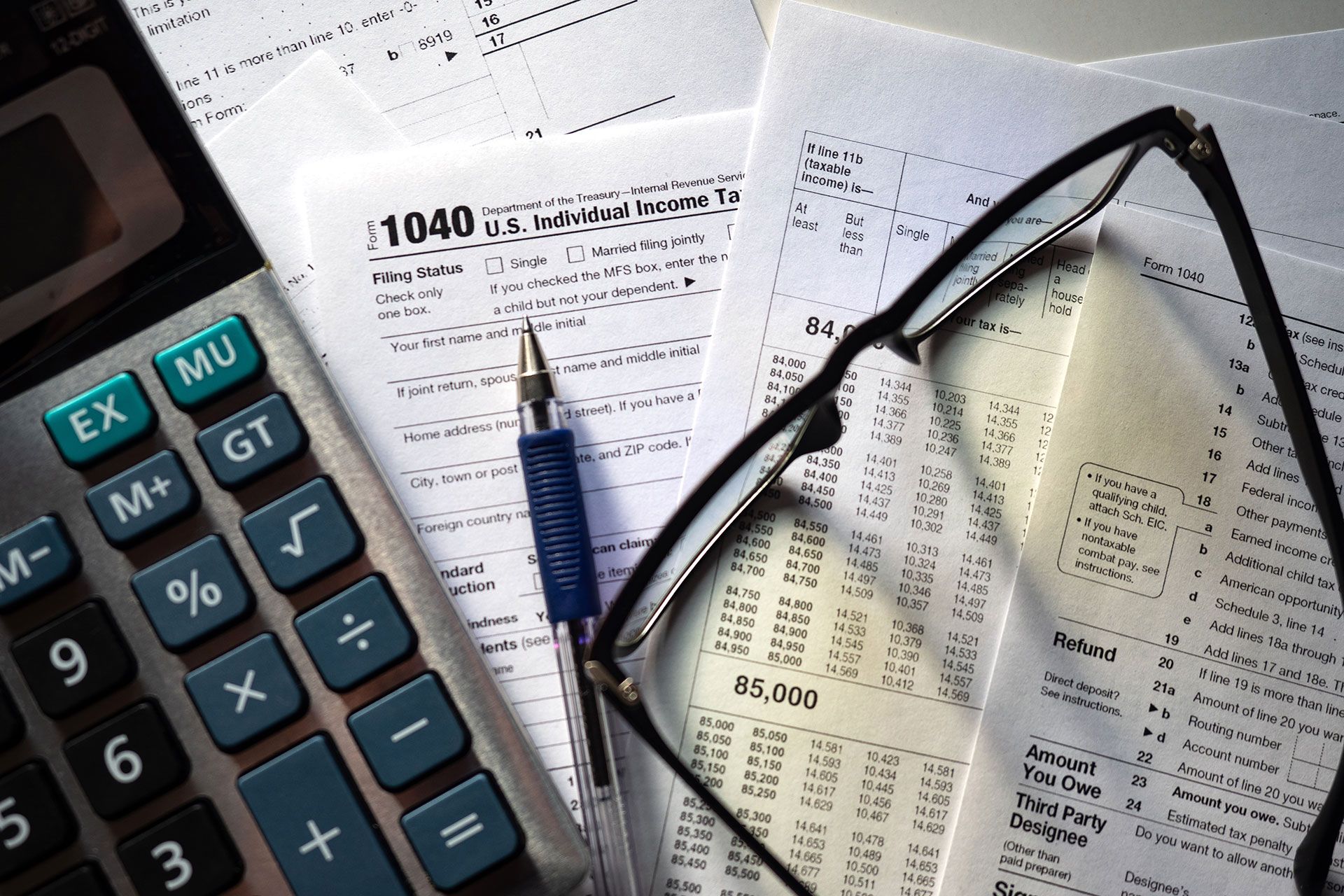 Calculator, tax forms, pen, and glasses on a desk, representing tax preparation.