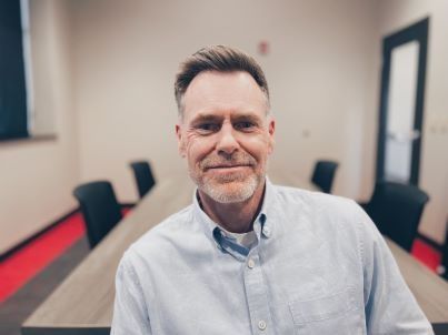 Man in blue shirt smiles in a conference room; long table, chairs, neutral walls, red carpet.