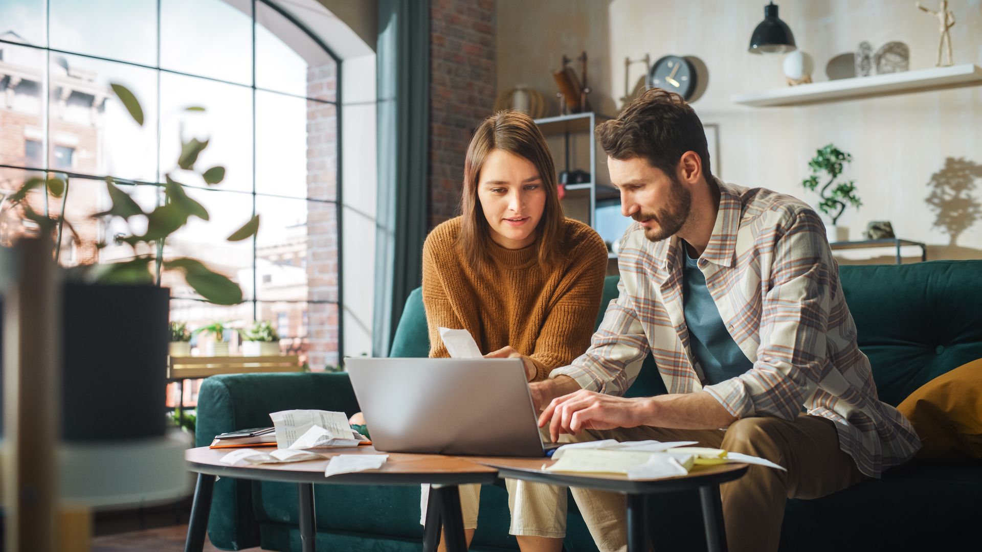 Couple on sofa looking at laptop, reviewing documents on coffee table. Bright living room.