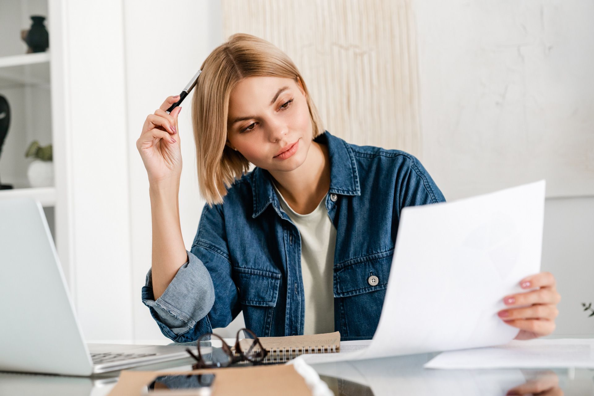 Woman in denim shirt reviews papers, pen in hand, at a desk with a laptop and calculator.