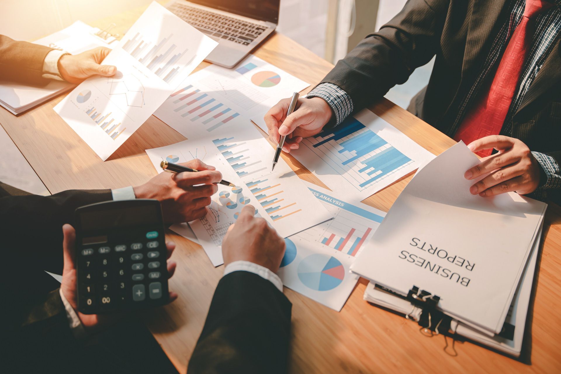 Businesspeople analyze financial reports around a table, using a laptop and calculator.