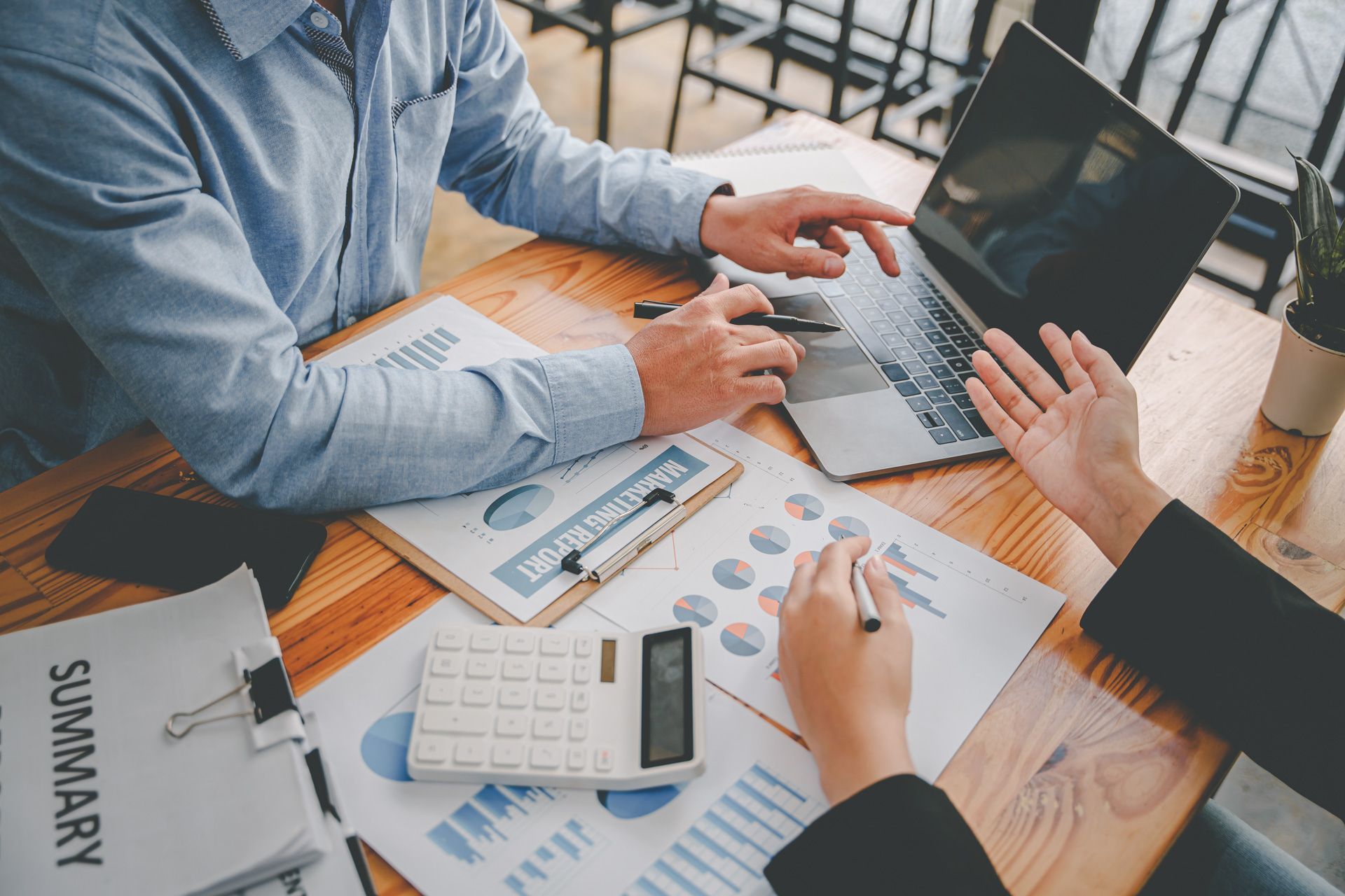 Two people review financial documents at a wooden table with a laptop and calculator.