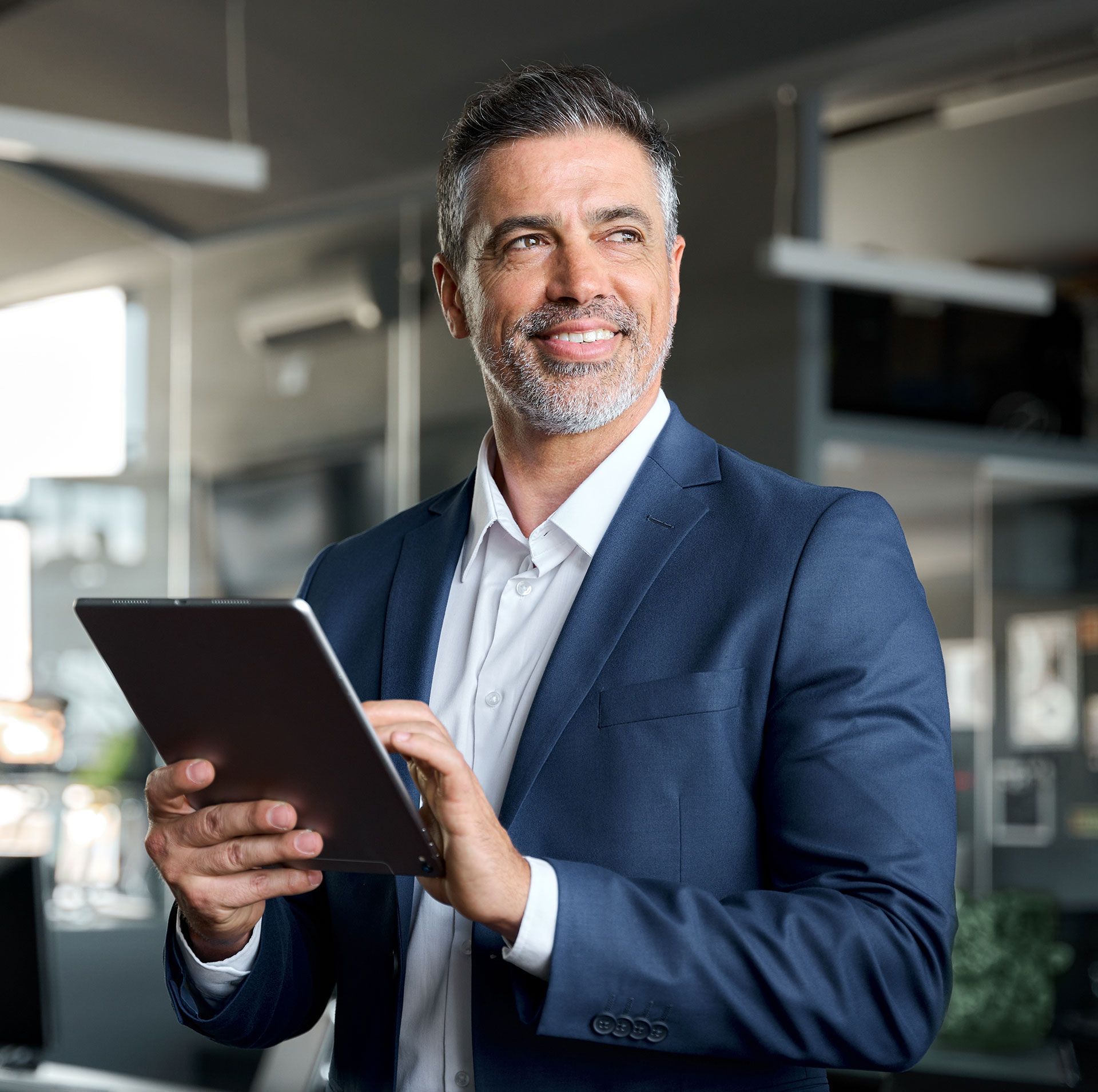 Man in suit holding a tablet, smiling, looking to the side in an office setting.