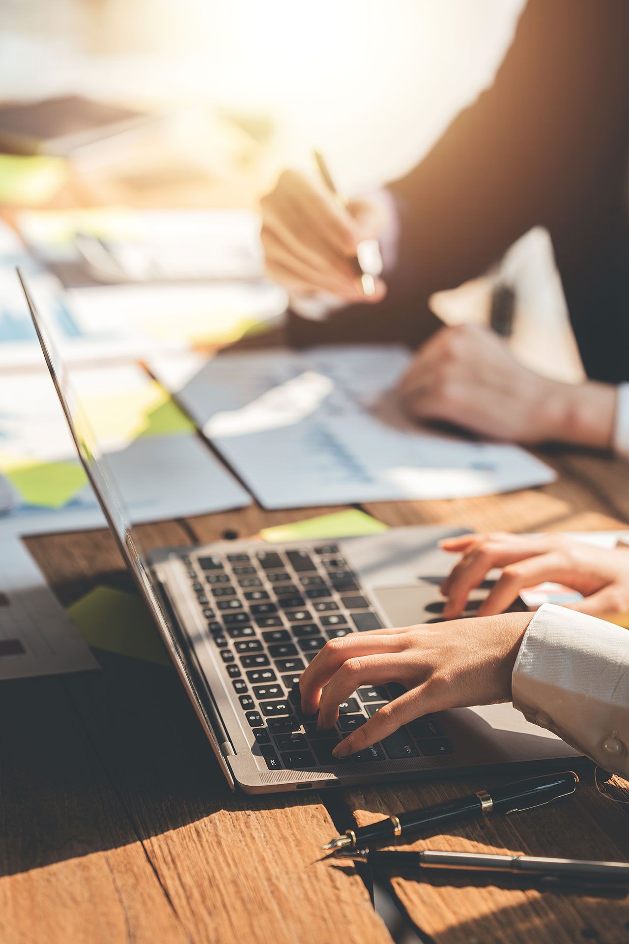 Hands typing on laptop at wooden desk with papers and pen. Another person in background.