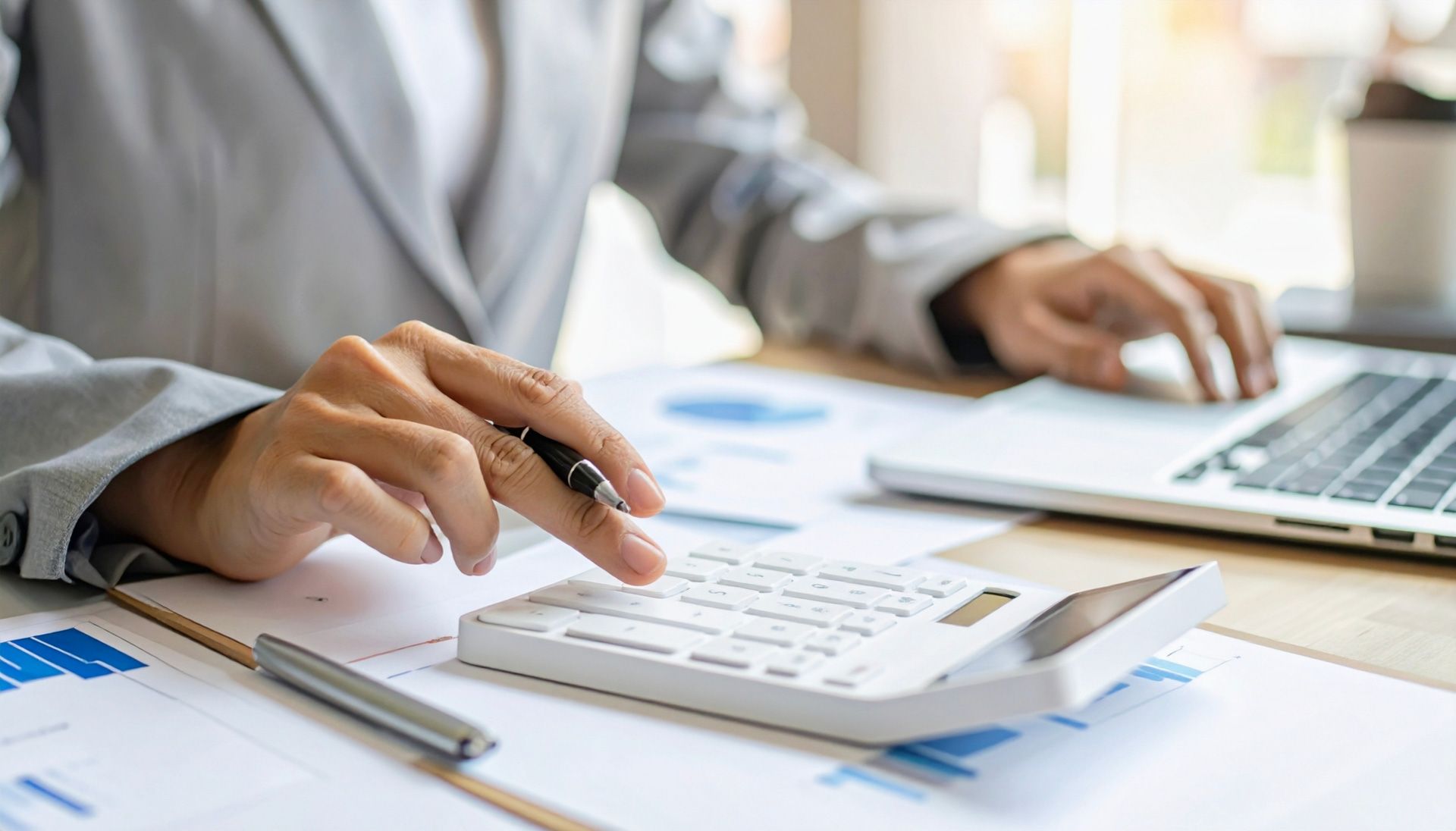 Hands calculating with a calculator, using a laptop, and surrounded by financial documents.