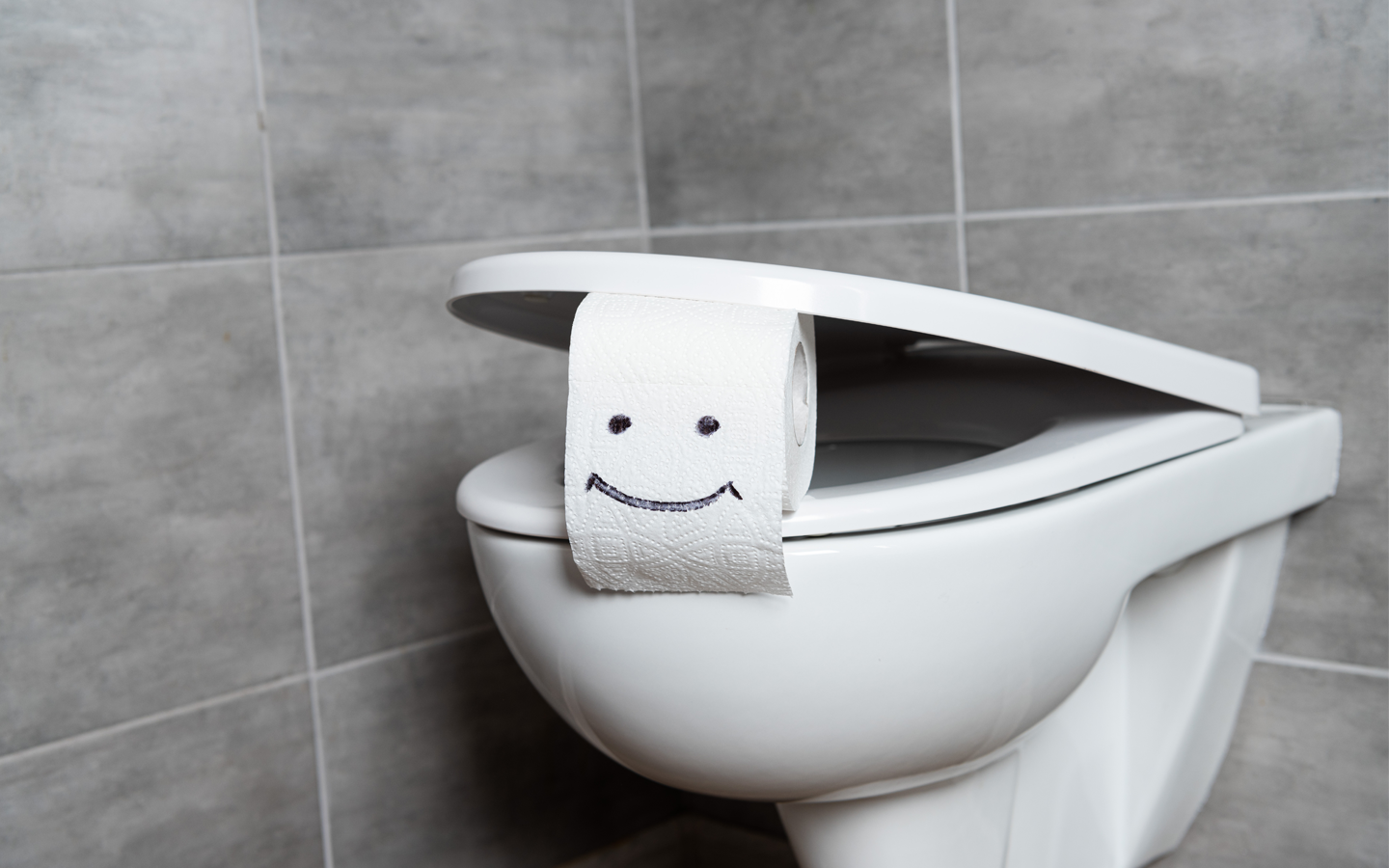 Smiling face drawn on toilet paper roll atop a white toilet in a tiled bathroom.