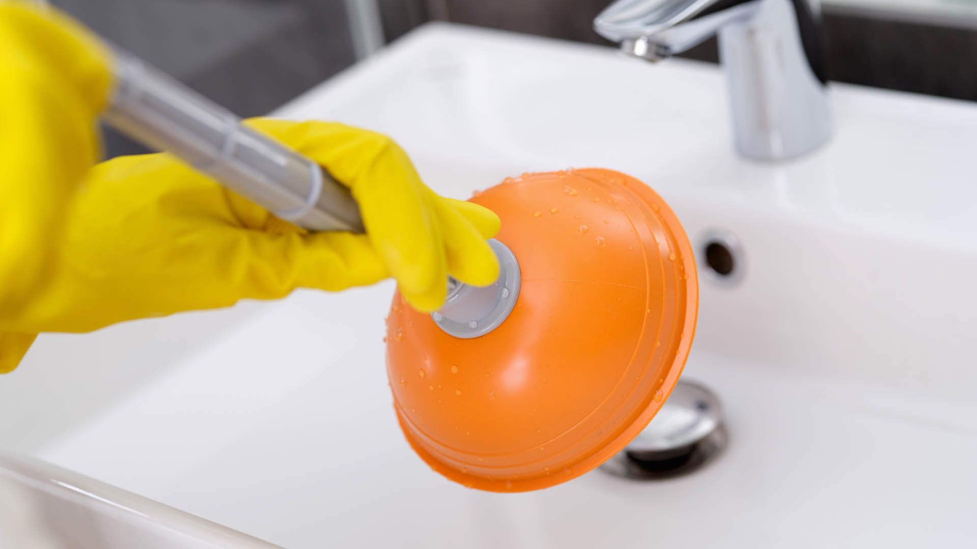 Person in yellow gloves using orange plunger on a white sink.