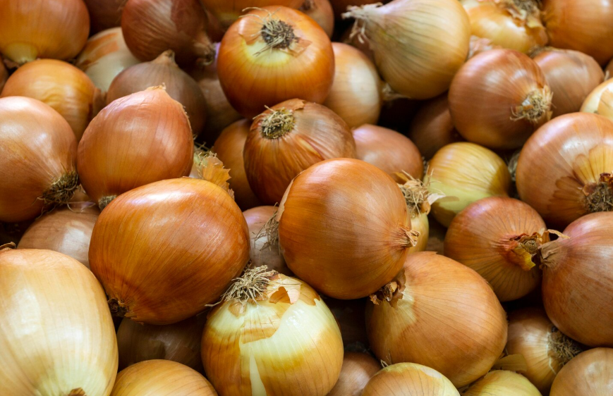 Montón de cebollas marrones, algunas con raíces visibles, muy juntas.